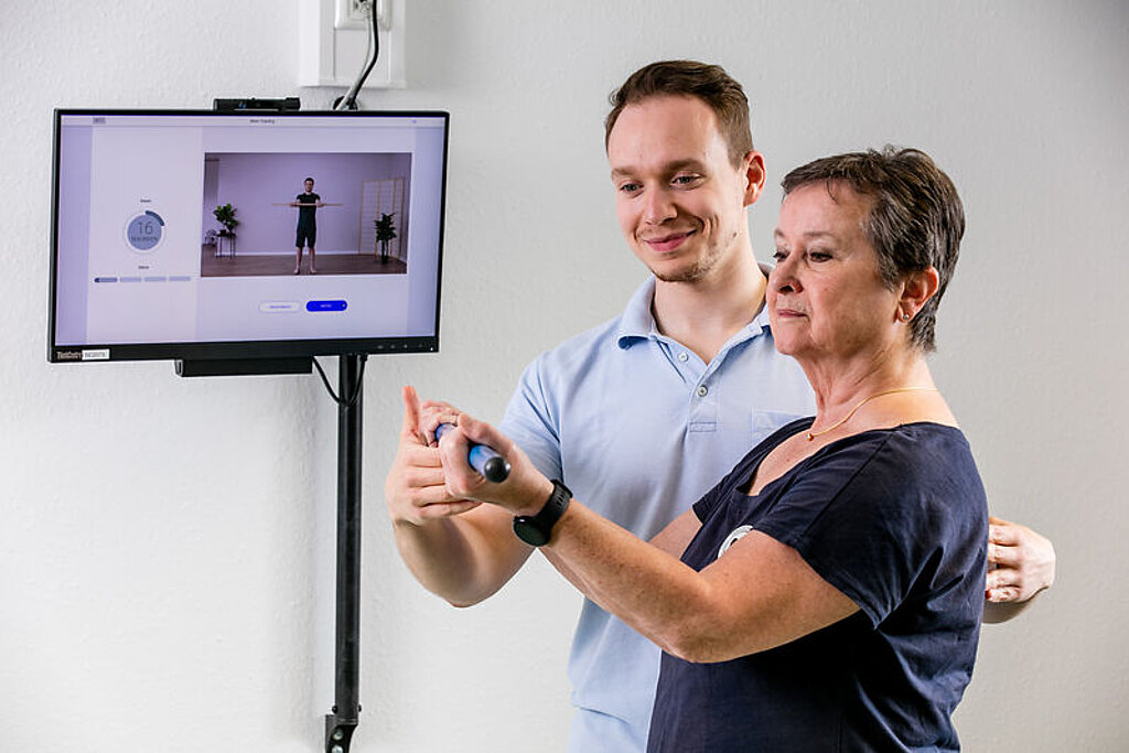 Ein Therapeut unterstützt eine Patientin in ihrem Training, welches sie anhand eines Trainingsvideos durchführt.