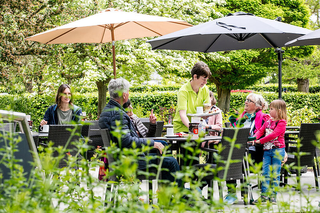 Auf einer Terrasse im Grünen sitzen Menschen an Tischen, eine Familie wird von einer Servicekraft bedient.